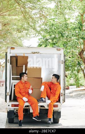 Tired movers sitting in loaded van trunk, drinking water and talking during short break Stock Photo