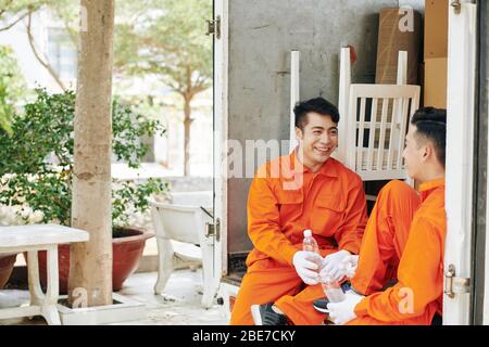 Cheerful young Vietnamese movers sitting in car trunk, drinking water and joking around after loading van with cardboard boxes Stock Photo