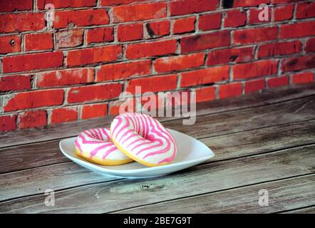Two fresh donuts in pink and white glaze on a ceramic plate stand on a wooden table against a brick wall. Close-up. Stock Photo