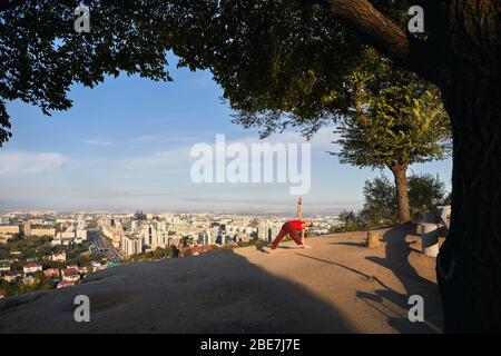 Fit man in red costume doing yoga triangle asana in the park with city and mountain on background in Almaty, Kazakhstan Stock Photo