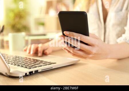 Close up of woman hands checking smart phone using laptop on a desk at home Stock Photo