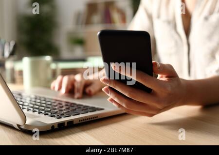 Close up of woman hands using laptop checking smart phone at night at home