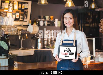 asian barista holding tabblet sign we’re open for takeaway orders only infront of counterbar.social distancing concept when coronavirus is outbreak in Stock Photo