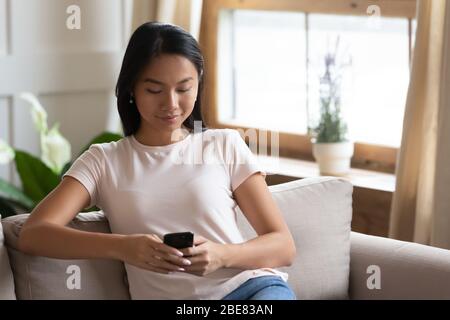 Asian woman sitting on couch using smartphone communicating with friends Stock Photo