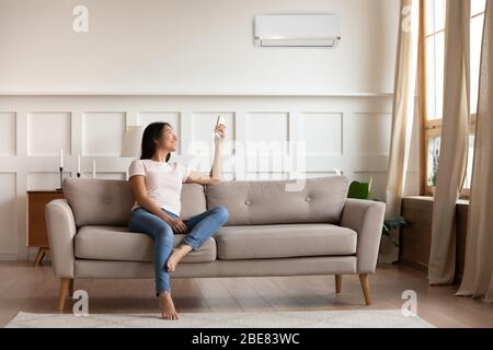 Asian woman switching on air conditioner while resting seated indoors Stock Photo