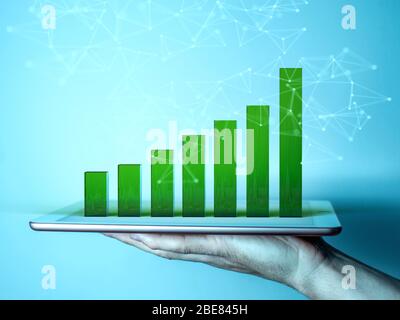 A man's hand holding a tablet. The screen of the gadget shows a 3d graph in the form of green columns illustrating growth Stock Photo