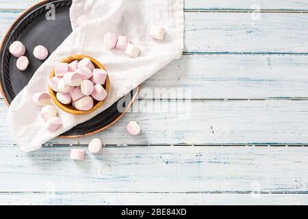 Top view of a wooden bowl full of pink and white marshmallows with some scattered around on a white table cloth, black tray and white wooden surface. Stock Photo
