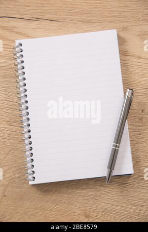 White lined notepad with a metal ring wire binding and a silver pen opened on a wooden surface. Stock Photo