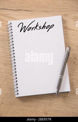 Blank spiral notebook with a Workshop headline and empty lines placed on a wooden table and a metallic silver pen positioned on top of it. Stock Photo