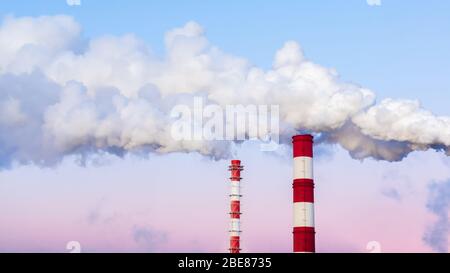 Striped chimneys emit thick white smoke in the evening blue pink sky. Air pollution, environmental problems. Tall pipes against the backdrop of a beau Stock Photo