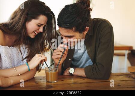 Happy young couple in love at cafe drinking a coffee from same glass. Young man and woman enjoying a coffee on table. Stock Photo