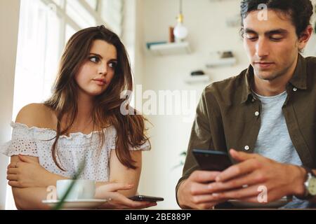 Woman looking unhappy while her man paying no attention to her and busy using his mobile phone. Sulking woman sitting next to man reading text message Stock Photo