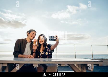 Woman taking selfie with smartphone with her boyfriend holding an ice cream sitting outdoors. Couple taking selfie on their date. Stock Photo