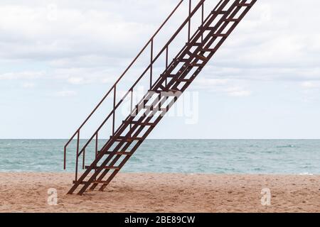 Rusty metal stairway on sandy beach under cloudy sky background Stock Photo