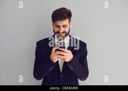 Young man in a jacket with a mobile in his hand typing sms smiles laughing joyful happy positive on a gray background. Businessman entrepreneur freela Stock Photo