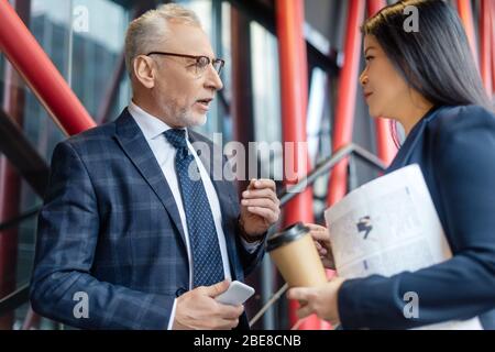 businessman with smartphone and asian businesswoman with paper cup talking and looking at each other Stock Photo