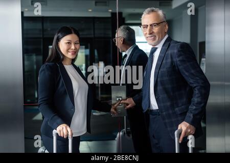 smiling businessman and asian businesswoman with travel bags pushing button of elevator Stock Photo