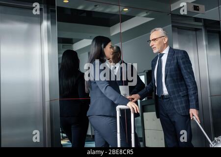 businessman pushing button of elevator and talking to asian businesswoman with travel bag Stock Photo