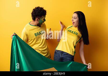 male african american football fan with painted face and happy girl gesturing and holding brazilian flag on yellow Stock Photo