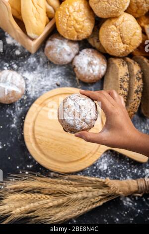 Top view the hand picks up the loaves of bread to put on an empty wooden cutting board and wheat on the table with many kinds of bread. Stock Photo