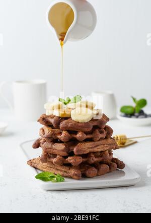 Chocolate banana waffles on white table, vertical. Sweet brunch, maple syrup flow Stock Photo