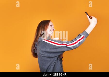 Cute playful young woman taking selfie with mobile phone on yellow background Stock Photo