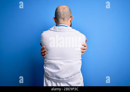 Handsome bald doctor man with beard wearing glasses and stethoscope over blue background Hugging oneself happy and positive from backwards. Self love Stock Photo