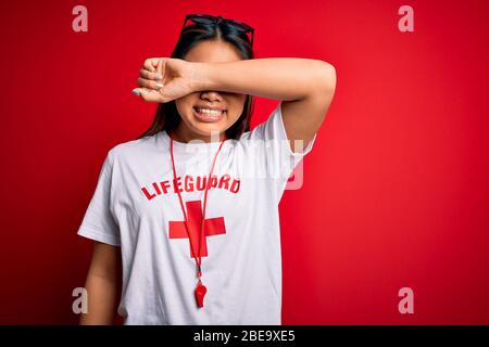 Young asian lifeguard girl wearing t-shirt with red cross using whistle over isolated background Smiling cheerful playing peek a boo with hands showin Stock Photo