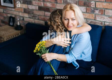 Beautiful blonde young woman and her curly charming little daughter are hugging at the home in cozy living room Stock Photo