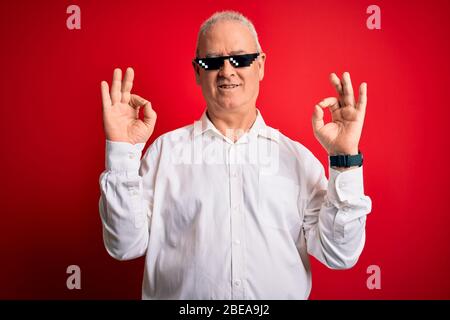 Middle age hoary man wearing funny sunglasses over isolated red background relax and smiling with eyes closed doing meditation gesture with fingers. Y Stock Photo