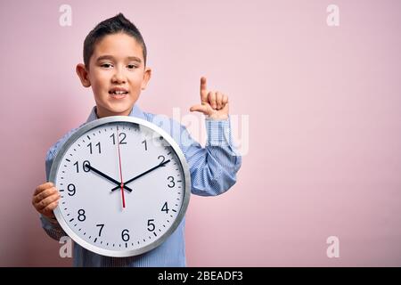 Young little boy kid holding big minute clock over isolated pink background surprised with an idea or question pointing finger with happy face, number Stock Photo