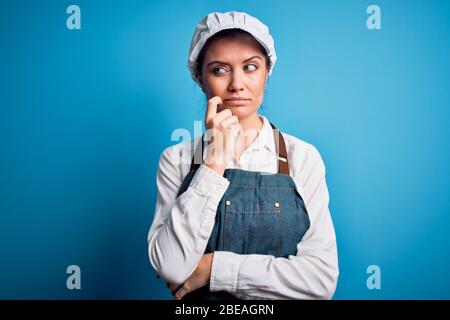 Young beautiful baker woman with blue eyes wearing apron and cap over blue background with hand on chin thinking about question, pensive expression. S Stock Photo