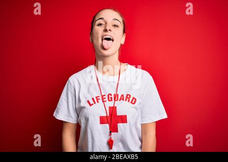 Beautiful lifeguard woman wearing t-shirt with red cross using whistle over isolated background sticking tongue out happy with funny expression. Emoti Stock Photo