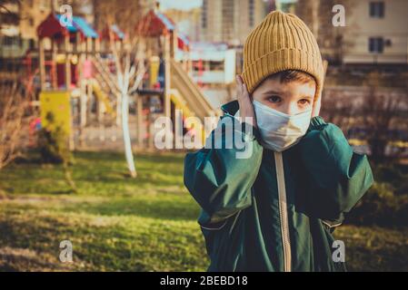 A boy in a protective mask stands near a playground in the street during the pandemic of coronavirus and Covid - 19