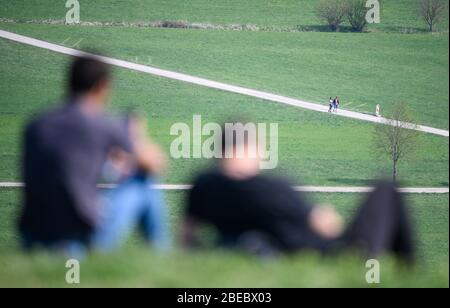 13 April 2020, Baden-Wuerttemberg, Stuttgart: Two men are sitting on a hill in the sun. In the background two walkers are walking. To slow down the spread of the corona virus, a contact ban is in place. Photo: Sebastian Gollnow/dpa Stock Photo