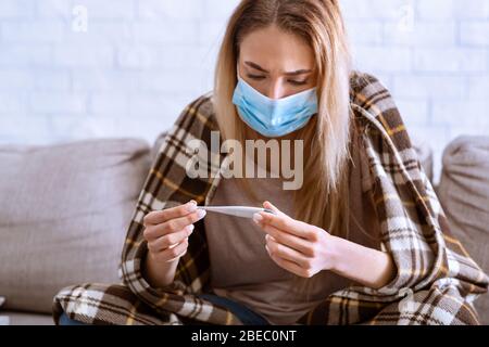 Woman in mask, covered blanket looks at thermometer Stock Photo