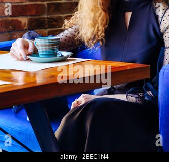 A young lady with curly brown hair is resting in the cafeteria at a table by the window. We see the table itself and the cup held by the girl's hand, her other hand resting on her lap, and the light from the window falls on all this. Stock Photo