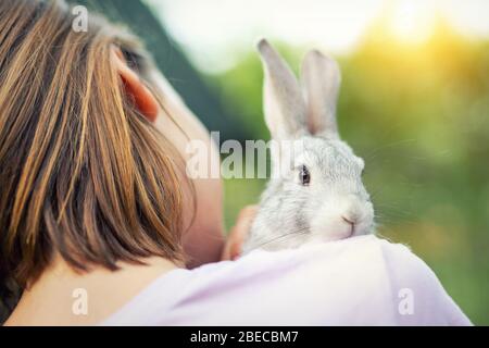Cute adorable sweet little rabbit sitting on shoulder of young adult brunette woman at warm sunset time country outdoors. Animal friendship and care Stock Photo