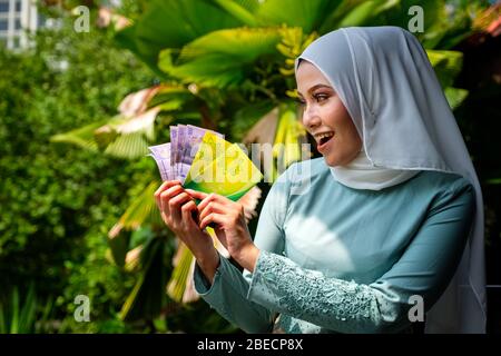 A portrait of young malay woman with surprise happy expression, holding an envelope of pocket money or raya angpao of Malaysian Currency. Translation Stock Photo