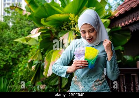 A portrait of young malay woman with surprise happy expression, holding an envelope of pocket money or raya angpao of Malaysian Currency. Translation Stock Photo