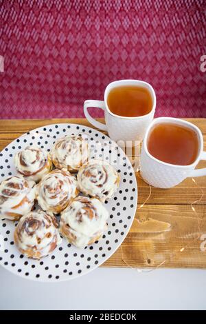 Two cups of black tea stand on a wooden tray. Fresh and fragrant cinnamon rolls close up lie on a plate with polka dots, beautiful morning. Close-up. Stock Photo