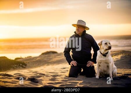 Middle-aged man kneeling next to his faithful dog on the beach at sunset Stock Photo