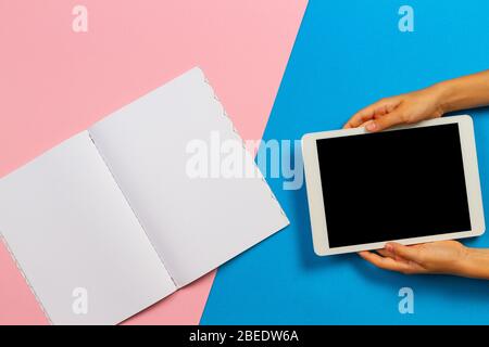 Kid hands with digital tablet computer and open paper notebook on light blue and pink background. Top view Stock Photo