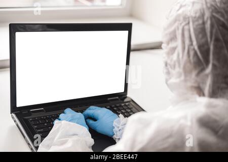 Person in protective white translucent suit, blue rubber gloves, medical mask sits at table at home and works or studies on laptop during quarantine. Stock Photo