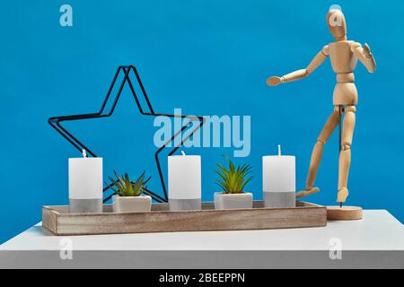 White table, figurine of human, green plants in pots, three big candles and one small in candlestick in form of iron star. Blue background. Close up Stock Photo