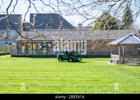 A man on a ride on  lawnmover cutting the grass of a primary school field  while the children are not at school Stock Photo