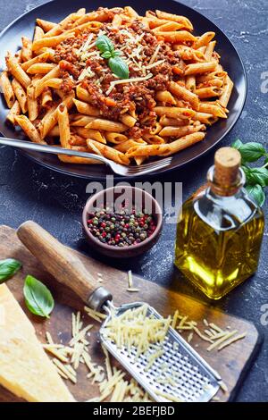 wholewheat pasta penne bolognese topped with shredded parmesan and basil on a black plate on a concrete table, vertical view from above