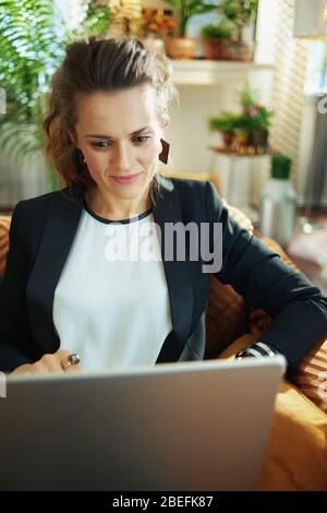 young housewife in white blouse and black jacket at modern home in sunny day reading on a laptop while sitting on couch. Stock Photo