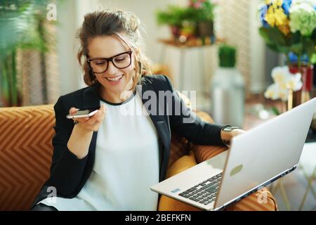 smiling stylish 40 years old housewife in white blouse and black jacket with laptop speaking on a mobile phone in the modern house in sunny day. Stock Photo