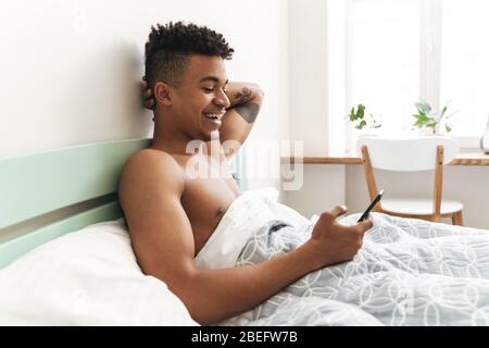 Image of optimistic pleased happy young african guy in the morning in bedroom using mobile phone in bed. Stock Photo